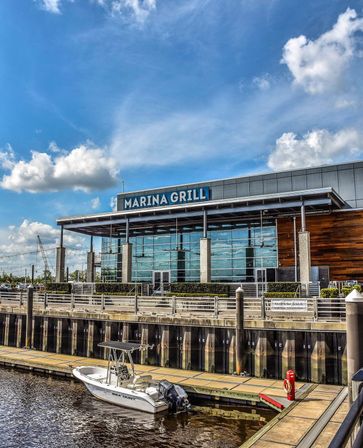 Sunlit waterfront grill with glass façade and covered patio, wooden dock and a small white motorboat moored in the marina under a bright blue sky with fluffy clouds.