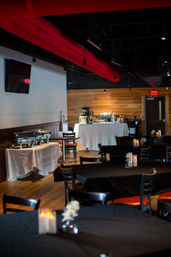 Industrial-chic restaurant event space with red exposed ductwork, wood-paneled bar, white-clothed buffet stations and black-tablecloth dining tables set with candles and small floral centerpieces.