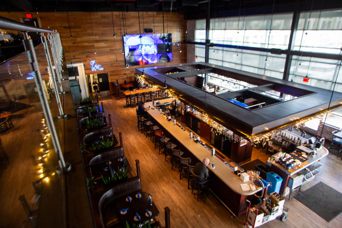 Elevated view of a modern industrial-style bar and restaurant interior with a long central island bar, leather booths, wood floors and walls, floor-to-ceiling windows, hanging string lights and a large wall-mounted screen.