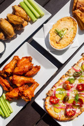 Outdoor appetizer spread: spicy buffalo chicken wings with celery and ranch, golden baked cheesy dip topped with scallions, and a vegetable-topped flatbread on white plates