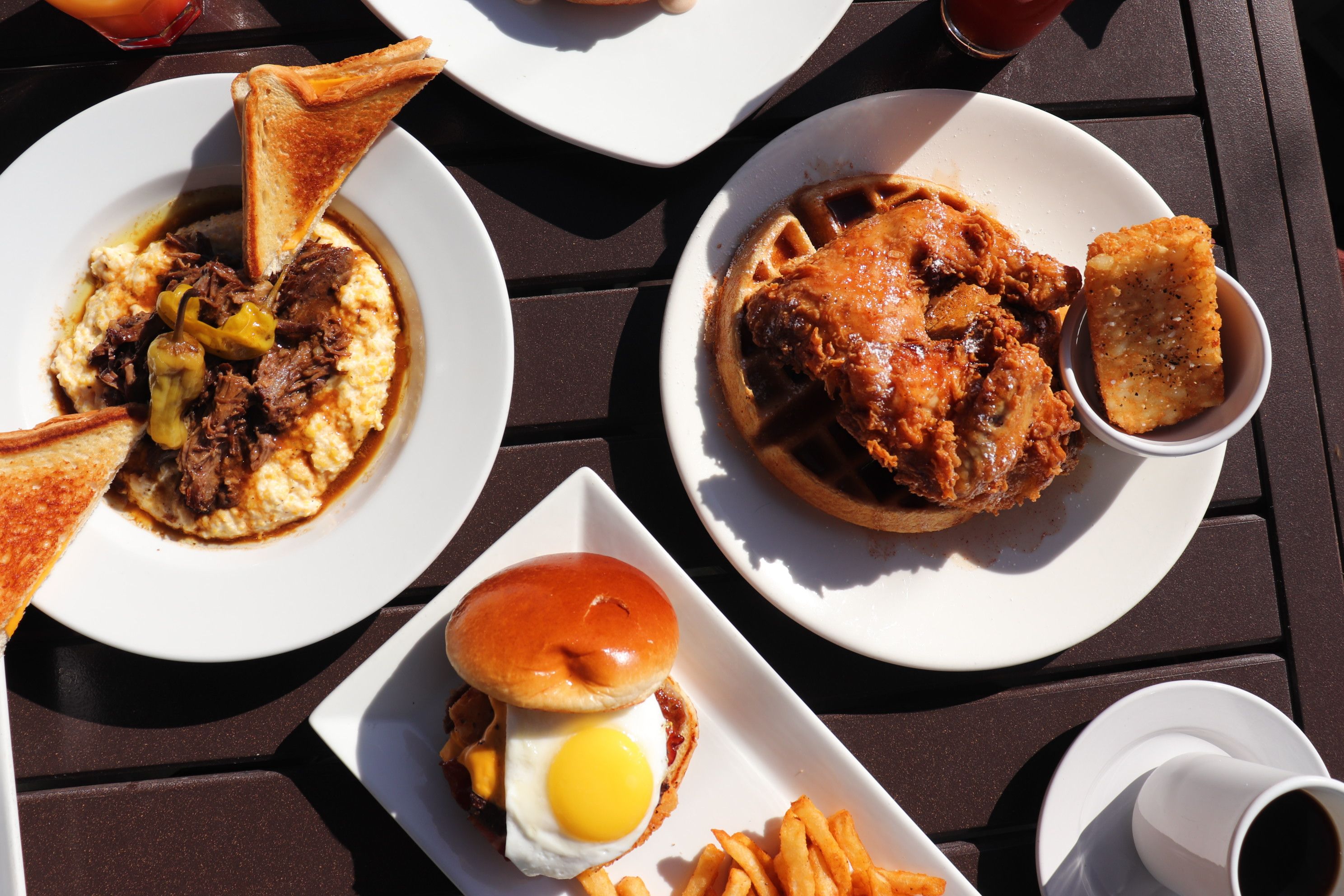 Southern-style outdoor brunch: crispy fried chicken on a waffle with syrup and hash brown, pulled beef over cheesy grits with toasted bread triangles, sunny-side egg breakfast sandwich with fries and coffee
