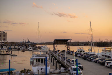 Marina at sunset with yachts and sailboats moored along a busy pier, parked cars lining the dock, a covered loading platform and string lights, calm water reflecting a pastel orange sky.