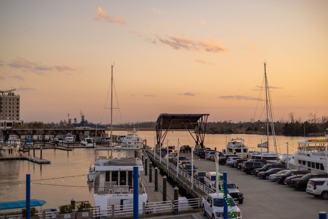 Marina at sunset with yachts and sailboats moored along a busy pier, parked cars lining the dock, a covered loading platform and string lights, calm water reflecting a pastel orange sky.