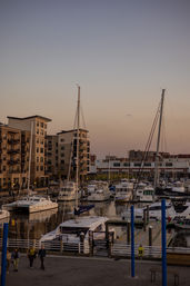 Urban waterfront marina at dusk with sailboats and yachts docked along wooden piers, modern apartment buildings and a boardwalk under a pastel sunset sky.