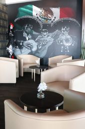 Day of the Dead–themed lounge with beige armchairs and dark round tables, small vases with white flowers, a cactus, and a mural of skeletal mariachis under a Mexican flag