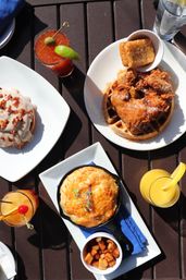 Sunny patio brunch spread: crispy fried chicken on a waffle with a hash brown, cheesy skillet biscuit with diced potatoes, glazed cinnamon roll, Bloody Mary and orange juice cocktails.