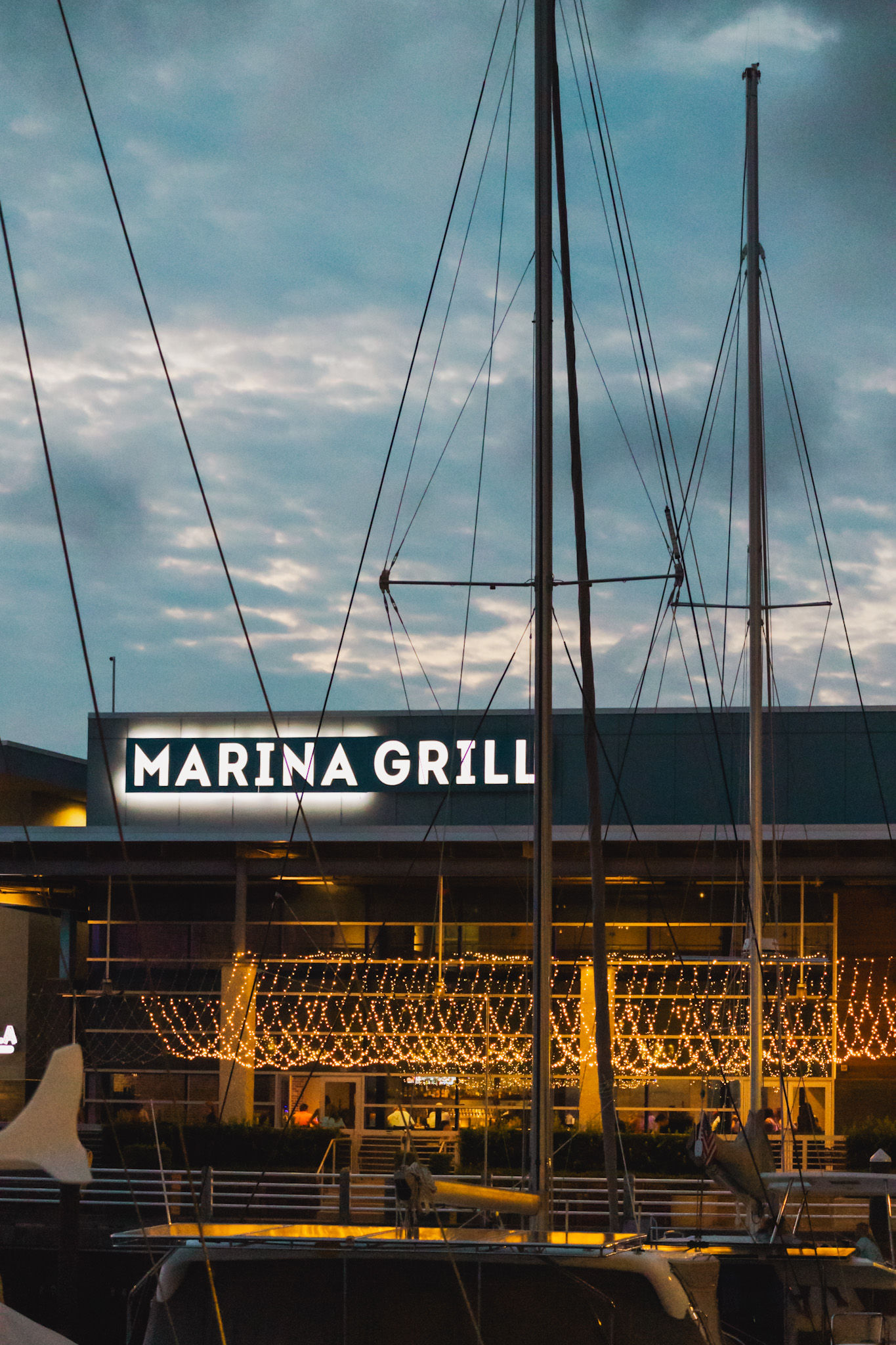 Docked sailboats and tall masts in a marina with a waterfront restaurant patio glowing with warm twinkling string lights at dusk
