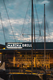 Docked sailboats and tall masts in a marina with a waterfront restaurant patio glowing with warm twinkling string lights at dusk