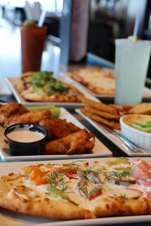 Close-up of cheesy flatbread topped with herbs and vegetables, fried chicken wings with ranch dip, pita chips and dips, and tall cocktails on a casual restaurant table
