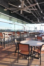 Sunny waterfront patio dining area with round wood-top tables, metal-framed chairs and high bar stools, oversized ceiling fans and a glass wall reflecting the marina.