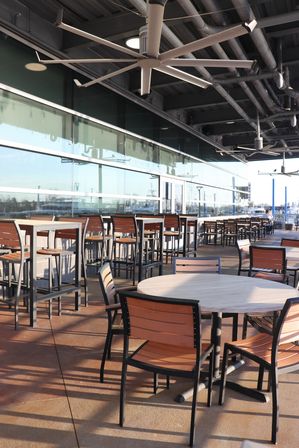 Sunny waterfront patio dining area with round wood-top tables, metal-framed chairs and high bar stools, oversized ceiling fans and a glass wall reflecting the marina.