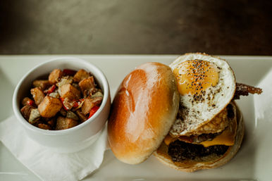 Brunch burger on a shiny brioche bun topped with a sunny-side-up egg, melted cheddar and crispy bacon, paired with a bowl of seasoned home fries and red peppers on a white plate.