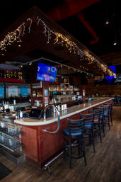 Modern bar interior with a long wood counter, row of high stools, back bar of liquor bottles and draft taps, hanging string lights and wall TVs showing sports, empty wood-floor dining area.