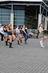 Energetic outdoor line-dance class in a downtown plaza — women in cowboy boots and casual summer outfits dancing in front of a modern glass building.