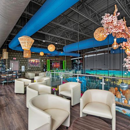 Mezzanine lounge with cream leather club chairs and round wood tables, glass railing overlooking a colorful tropical mural, bright blue exposed ceiling ducts and woven rattan pendant lights.
