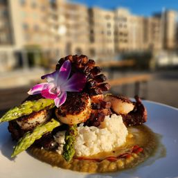 Seared scallops and grilled octopus tentacle atop white rice with asparagus, savory puree and a bright purple orchid garnish, served on an outdoor urban patio with blurred city buildings in the background.