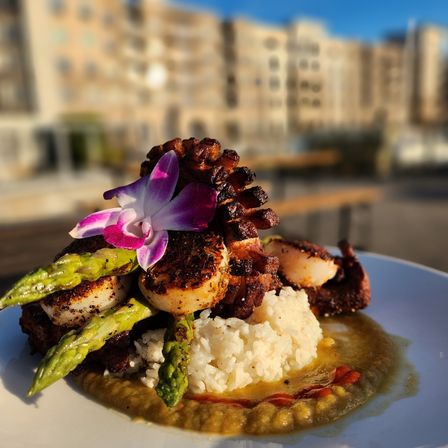 Seared scallops and grilled octopus tentacle atop white rice with asparagus, savory puree and a bright purple orchid garnish, served on an outdoor urban patio with blurred city buildings in the background.