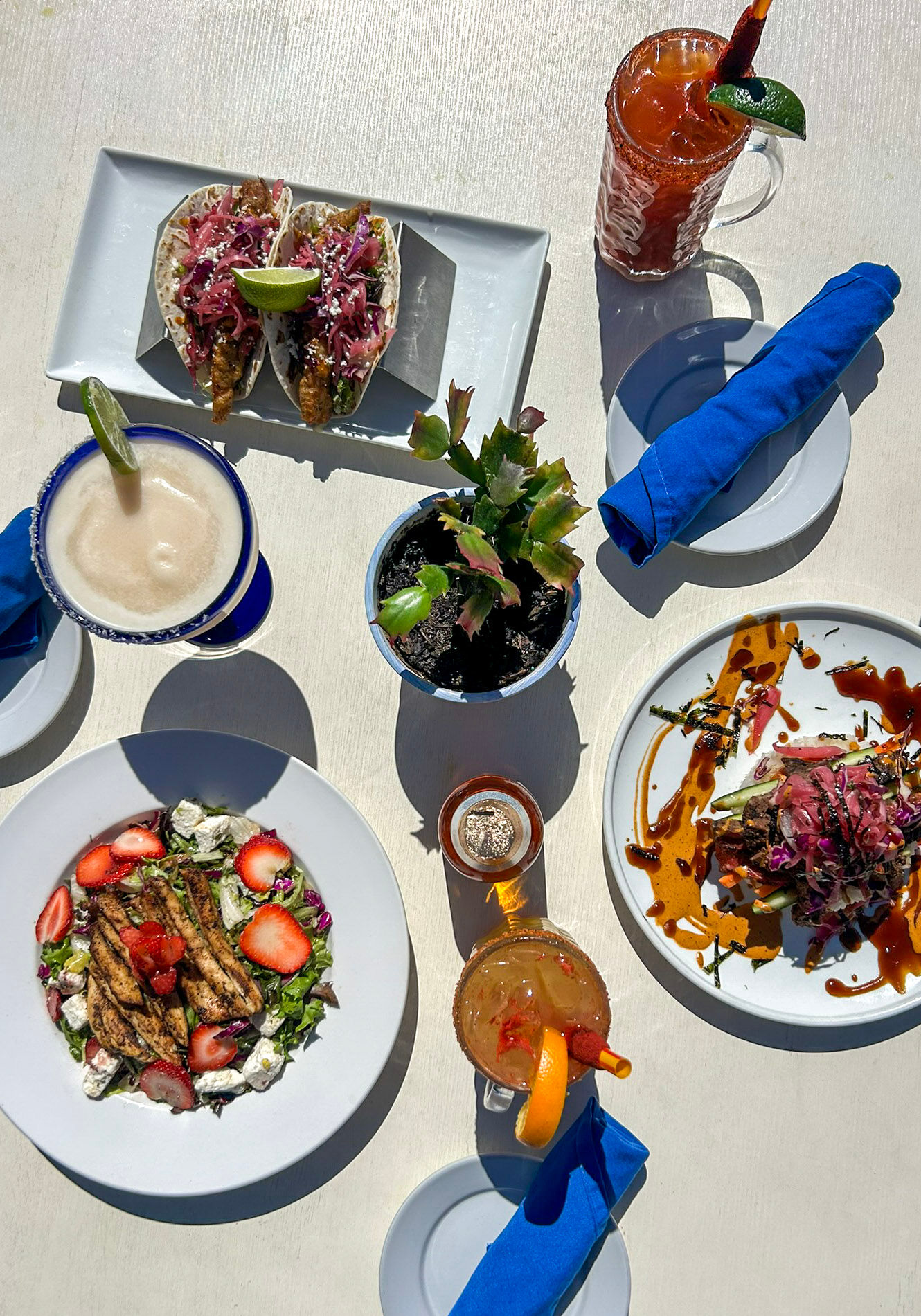 Top-down sunny patio view of a colorful brunch spread: two crispy tacos with lime, a salad topped with grilled protein and strawberries, a saucy composed plate with pickled slaw, a frozen margarita and two iced cocktails, small potted plant and bright blue napkins.