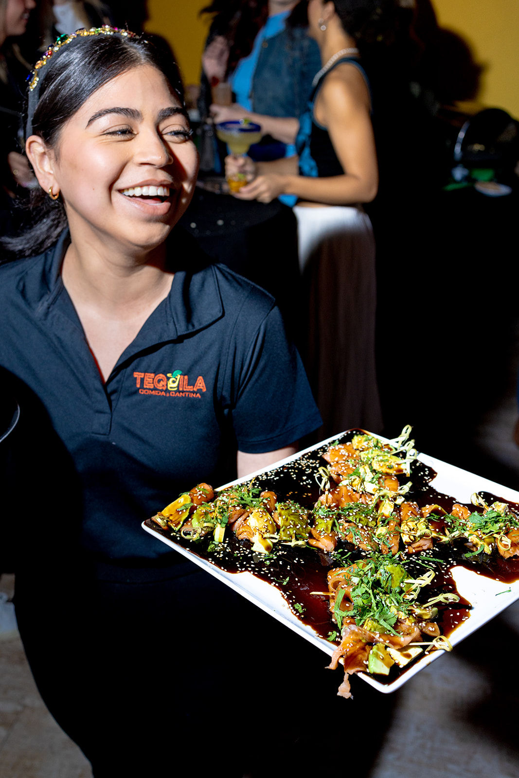 Smiling server in a black polo holding a white platter of glazed bite-size sushi appetizers garnished with sesame seeds and herbs at a lively indoor cocktail party