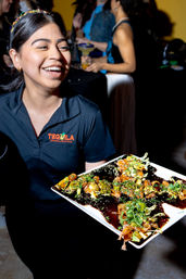 Smiling server in a black polo holding a white platter of glazed bite-size sushi appetizers garnished with sesame seeds and herbs at a lively indoor cocktail party