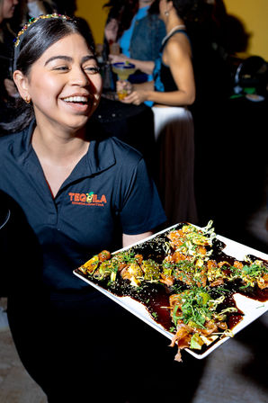 Smiling server in a black polo holding a white platter of glazed bite-size sushi appetizers garnished with sesame seeds and herbs at a lively indoor cocktail party