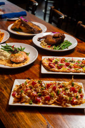 Assorted colorful dishes on a rustic wooden table at a casual restaurant: loaded crispy nachos with diced seafood and avocado, tomato-and-pickled-onion flatbread, grilled salmon with mashed cauliflower and green beans, and roasted chicken and steak plates.