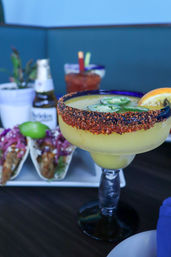 Frozen yellow margarita in a goblet with a zesty chili-lime rim, jalapeño slices and orange wedge, with tacos and a beer blurred in the background at a Mexican-style eatery