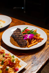 Grilled strip steak with peppercorn sauce, seasoned fries, sautéed green beans and red pepper slices, garnished with a purple orchid on a white plate on a wooden restaurant table.
