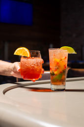 Two colorful iced cocktails on a bar counter — a short orange-garnished spritz held by a hand and a tall muddled-fruit highball with lime