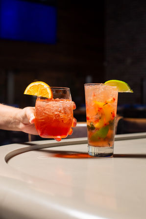 Two colorful iced cocktails on a bar counter — a short orange-garnished spritz held by a hand and a tall muddled-fruit highball with lime