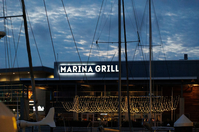 Dusk waterfront restaurant at a marina with sailboat masts in the foreground, a glowing sign and twinkling string lights over the outdoor terrace.