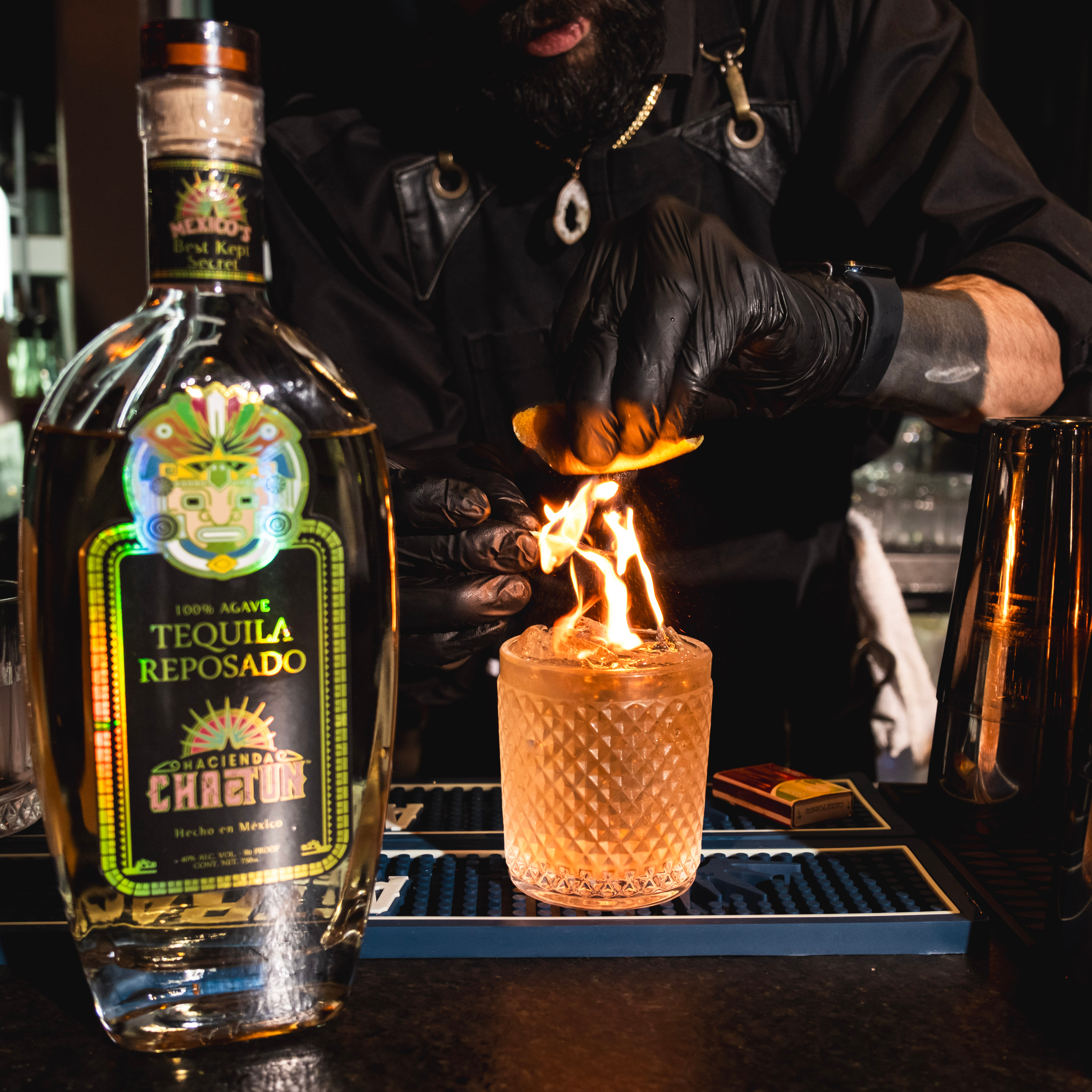 Cocktail bar scene: bartender flames orange zest over a tequila reposado cocktail in a textured glass, with a large tequila bottle in the foreground.