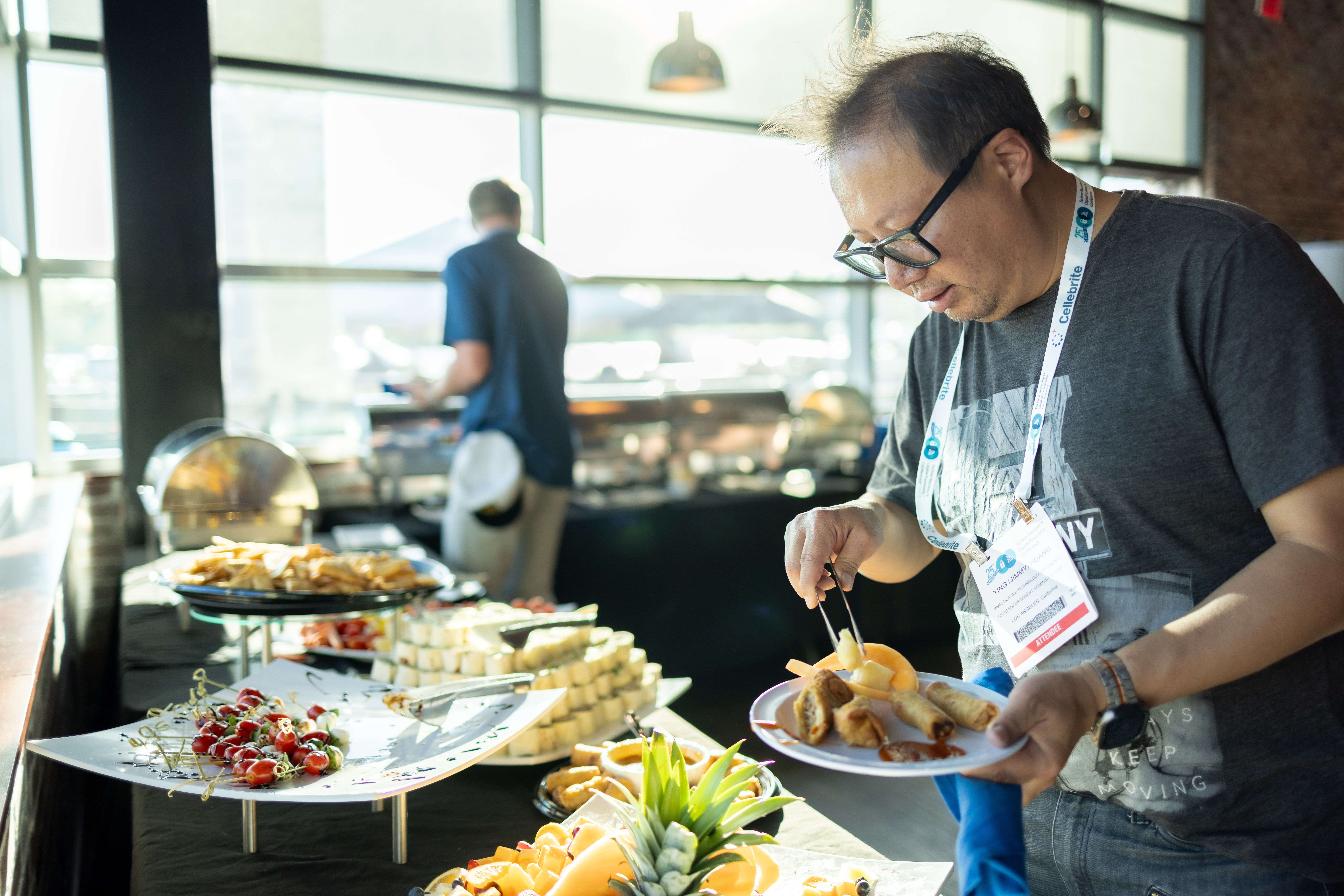 Conference attendee with lanyard filling a plate at an indoor buffet of spring rolls, fruit, cheese cubes and chips with a pineapple garnish.