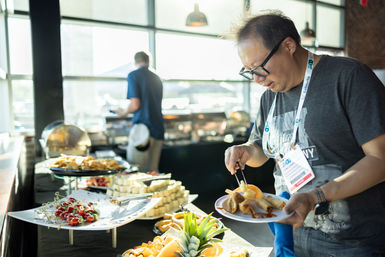 Conference attendee with lanyard filling a plate at an indoor buffet of spring rolls, fruit, cheese cubes and chips with a pineapple garnish.