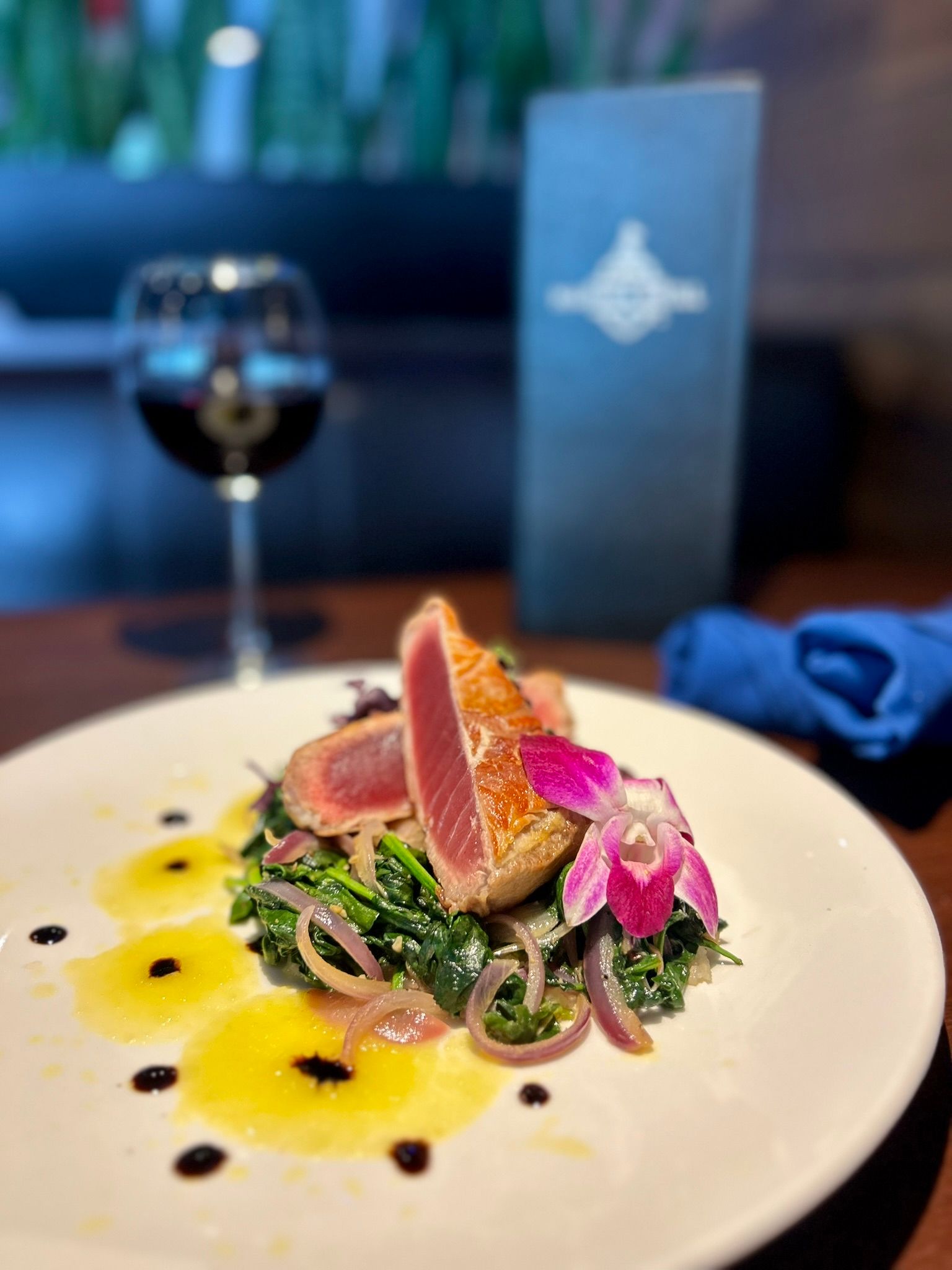 Seared ahi tuna slices on sautéed greens with pickled red onions, pink edible orchid and citrus oil dots on a white plate, wine glass and blurred restaurant menu in the background.