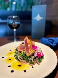 Seared ahi tuna slices on sautéed greens with pickled red onions, pink edible orchid and citrus oil dots on a white plate, wine glass and blurred restaurant menu in the background.