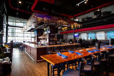 Spacious downtown industrial-style bar and restaurant interior with a central wooden bar, long communal table set with blue napkins, high ceilings, exposed red ducts, large windows and twinkling string lights.