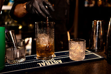 Gloved bartender hand pouring dark spirit into a tall mixing glass over ice, with a jigger, textured rocks glass and cocktail tools on a dim bar mat — cocktail prep, nightlife scene.