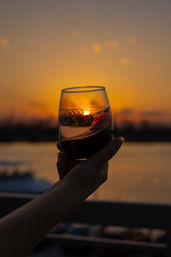 Wine glass held up to capture a golden coastal sunset over calm water — warm orange sky, silhouetted arm and sparkling reflections.