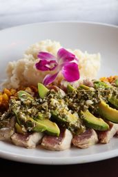 Seared ahi tuna slices topped with avocado and herb-garlic relish, served with white rice, roasted corn and a vibrant purple orchid on a white plate