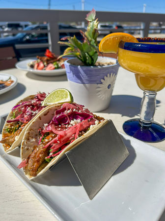 Sunny outdoor patio scene with two fish tacos in soft tortillas topped with pickled red cabbage, crumbled queso fresco and a lime wedge in a metal holder, beside a bright orange margarita garnished with an orange slice and a small potted succulent.