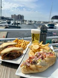Sunlit waterfront lunch at a marina: roast beef melt and a deli sandwich with fries and chips, plus a frothy beer on a dock with boats and yachts in the background.
