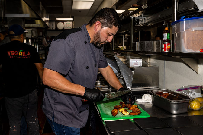 Busy restaurant kitchen: chef with gloved hands slicing caramelized fried plantains on a green cutting board at a stainless steel prep station.