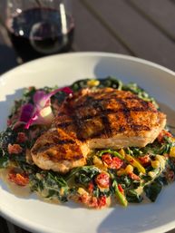 Grilled pork chop with char marks on creamy sautéed spinach and sun-dried tomatoes, garnished with an edible flower and a glass of red wine on a white plate outdoors.