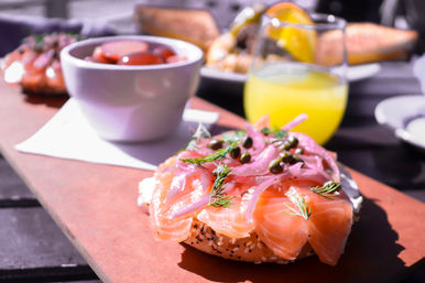 Sunlit patio brunch: sesame bagel with cream cheese topped with thinly sliced smoked salmon (lox), pickled red onions, capers and dill, with a bowl of cherry tomatoes and a glass of orange juice in the background.
