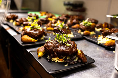 Mole-glazed braised beef on rice and black beans with fried plantains and microgreen garnish, plated on black square dishes lined up in a busy restaurant kitchen
