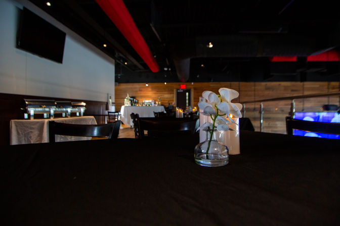 Modern restaurant event space interior with wood-paneled walls and exposed red ductwork, black-tablecloth table topped with a glass vase of white orchids and pillar candles, buffet and bar in the background.