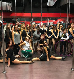 Energetic pole-fitness class group photo in a mirrored dance studio, performers in cabaret-style costumes and workout wear, two women doing front splits in the foreground with chrome poles and a purple hula hoop visible.