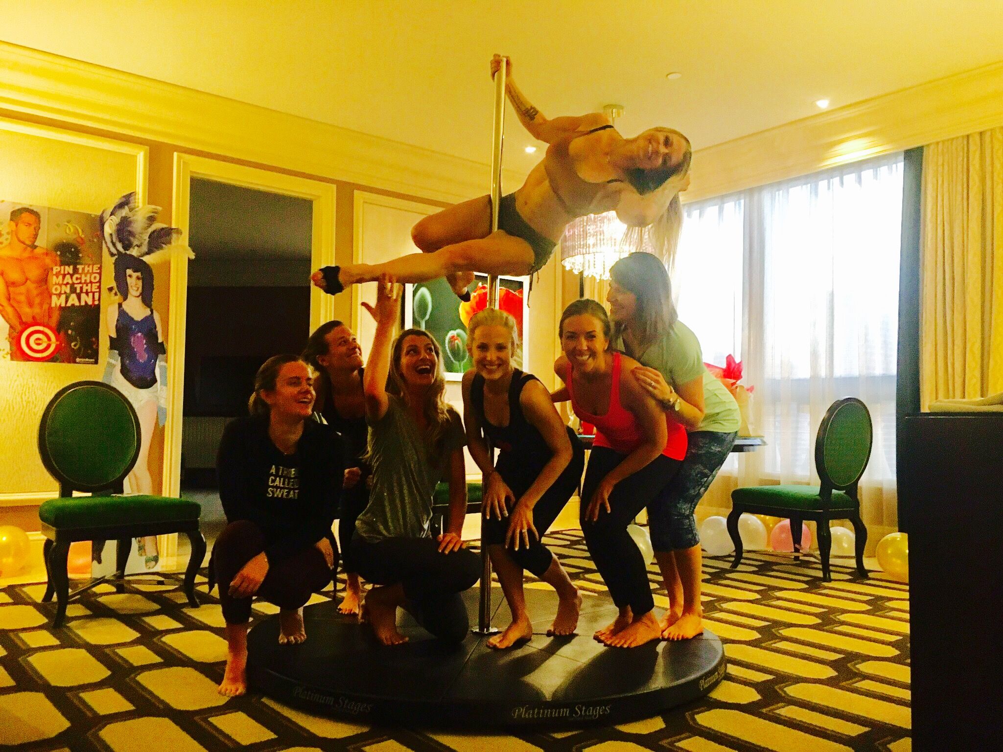 Group of seven women in a hotel suite pole-dancing class, one performing a horizontal pole move above a portable stage while friends cheer, with balloons and chairs in the background.