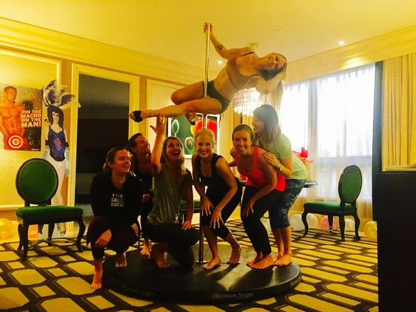 Group of seven women in a hotel suite pole-dancing class, one performing a horizontal pole move above a portable stage while friends cheer, with balloons and chairs in the background.